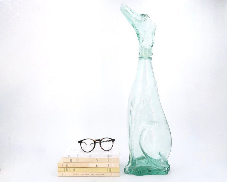 Peut inclure: Une bouteille en verre vert clair avec un design abstrait unique. &Agrave; c&ocirc;t&eacute;, une pile de livres et des lunettes rondes &agrave; monture noire. Le design de la bouteille ressemble &agrave; un oiseau ou un animal.