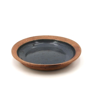 Op de afbeelding: Een rond keramieken bord met een donkerblauwe, glanzende en gespikkelde binnenkant, gecontrasteerd door een brede, ongeglazuurde en getextureerde terracotta-kleurige rand met fijne donkerbruine spikkels.
