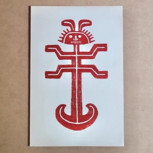 May include: A red and white print of a stylized figure with a sun-like face, rabbit ears, and a body resembling a cross. The figure is printed on a white rectangular paper.