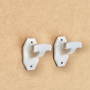 May include: Two white ceramic wall hooks, each featuring a T-shaped design with a smooth, glossy finish. Each hook has an elongated hexagonal base plate with two small circular screw holes, one at the top and one at the bottom for mounting.