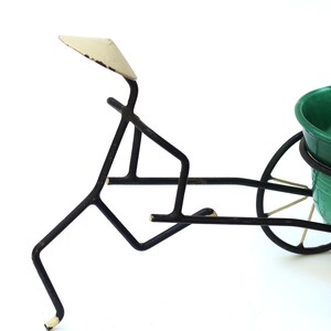 Op de afbeelding: Een grillige plantenbak in vintage-stijl met een stokfiguur die een kruiwagen duwt. De kruiwagen bevat een groene keramische kom. Het metalen frame is zwart met gebroken witte accenten, wat een charmante, rustieke uitstraling creëert.