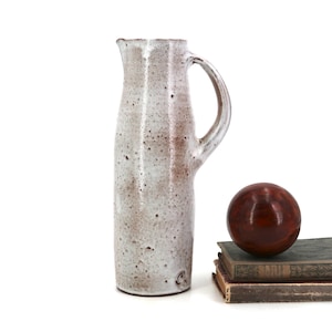 May include: A tall, cylindrical ceramic pitcher with a speckled off-white glaze and a curved handle. Beside it sits a dark reddish-brown wooden sphere resting on top of two stacked, weathered hardcover books with muted green and brown covers.