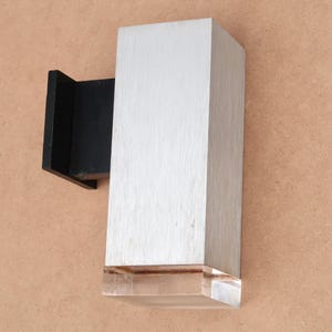 Könnte beinhalten: Eine moderne Wandleuchte mit gebürsteter Silberoberfläche und klarer, geometrischer Basis. Die Leuchte ist an einer schwarzen Halterung befestigt. Das Design ist minimalistisch und modern.