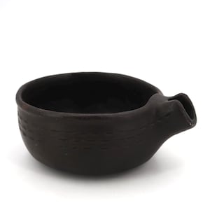 May include: A dark brown ceramic bowl with a rounded shape and a small pouring spout on the side. The exterior surface features a subtle, horizontal textured pattern consisting of dashed lines encircling the body of the vessel.