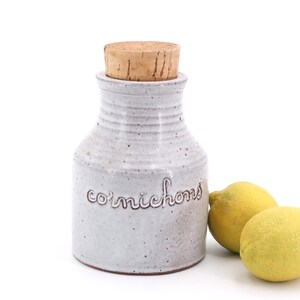 Puede incluir: Tarro de cerámica blanco con tapa de corcho y la palabra "cornichons" escrita en la parte delantera. Dos limones amarillos están en primer plano.