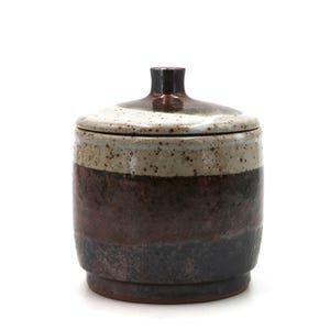 Puede incluir: Un tarro de cerámica con tapa, con un diseño en capas en tonos marrón, beige y negro. El tarro tiene forma cilíndrica con una base ligeramente más ancha y un pequeño pomo marrón oscuro en la tapa. La textura moteada añade interés visual.