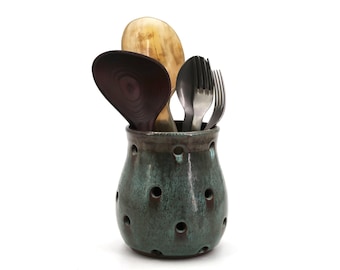 Peut inclure: Un pot à ustensiles en céramique turquoise avec de multiples ouvertures rondes, contenant des cuillères en bois et des fourchettes en métal. Le pot a un glaçage tacheté et un bord foncé. Les cuillères sont de différentes nuances de brun et de beige.