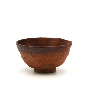 Può includere: Una piccola ciotola in ceramica di forma rotonda, modellata a mano, con una superficie ruvida e strutturata. L'esterno e l'interno presentano un profondo colore bruno-rossastro terroso con toni marroni più scuri e screziati sul bordo.