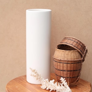 Puede incluir: Un jarrón cilíndrico blanco alto se encuentra junto a dos cestas tejidas y un trozo de coral blanco sobre una superficie de madera. Las cestas son marrones con rayas negras. El fondo es de color beige.