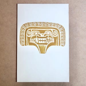May include: A gold-colored print on white paper featuring a stylized face with a wide grin and intricate details. The face has a rectangular shape with a decorative border and a prominent, toothy mouth.