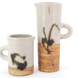 May include: Two ceramic mugs with brown and white glaze. The larger mug has a handle and a brown band around the bottom. The smaller mug has a handle and a brown band around the bottom.
