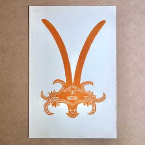 May include: Orange print on white paper featuring a stylized design. The design includes a central decorative element with curved horns or antennae extending upwards. The artwork has a symmetrical, ornamental quality.