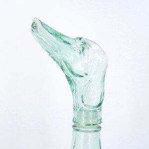 Peut inclure: Un bouchon de bouteille en verre transparent vert clair avec un design abstrait unique. Le bouchon pr&eacute;sente une forme incurv&eacute;e et allong&eacute;e avec des d&eacute;tails textur&eacute;s, ressemblant &agrave; une t&ecirc;te d'oiseau. La bouteille est partiellement visible, avec un col lisse et cylindrique.