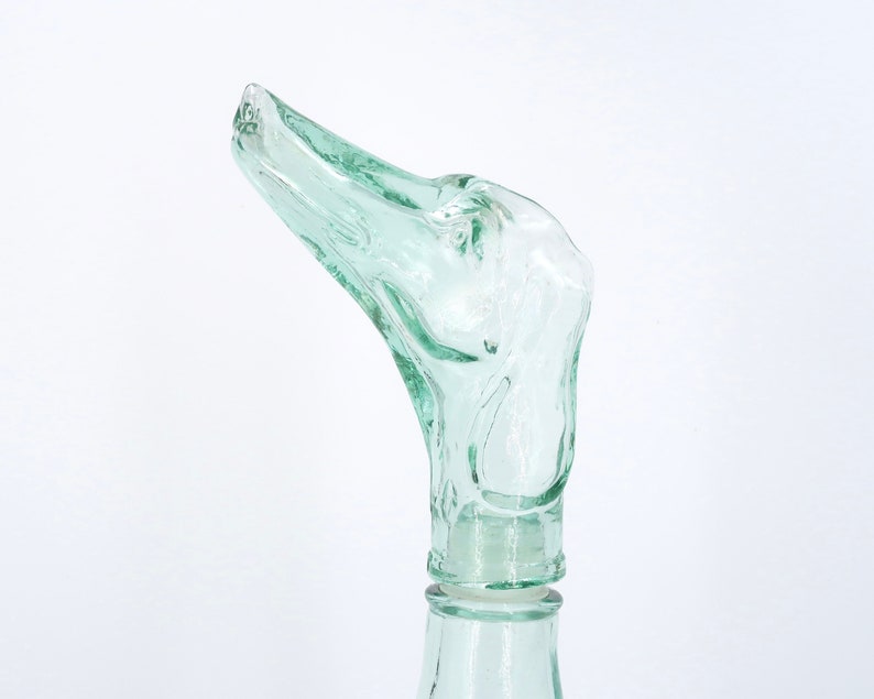Peut inclure: Un bouchon de bouteille en verre transparent vert clair avec un design abstrait unique. Le bouchon pr&eacute;sente une forme incurv&eacute;e et allong&eacute;e avec des d&eacute;tails textur&eacute;s, ressemblant &agrave; une t&ecirc;te d'oiseau. La bouteille est partiellement visible, avec un col lisse et cylindrique.
