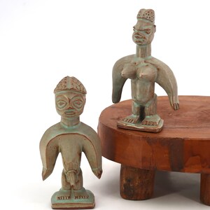May include: Two ceramic figurines with a teal glaze. One stands on a wooden pedestal. The figures have stylized features, including large eyes and outstretched arms. The base of one figure has text.