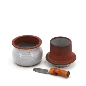 Peut inclure: Un beurrier en céramique avec un couvercle et un couteau à beurre. Le plat est blanc avec un bord marron, et le couvercle est marron. Le couteau a une lame argentée et un manche en bois. L'ensemble est sur fond blanc.