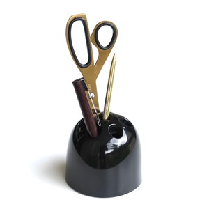May include: A black ceramic desk organizer with three compartments holds a pair of gold scissors, a gold pen, and a brown wooden pencil.