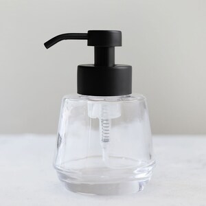 K&ouml;nnte beinhalten: Ein transparenter Seifenspender aus Glas mit einer schwarzen Pumpe. Der Spender hat ein strukturiertes, facettiertes Design.