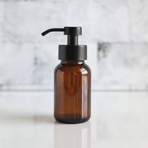 May include: A brown glass soap dispenser with a black pump. The dispenser has a cylindrical body and a rounded top. The background is a white marble-like surface.