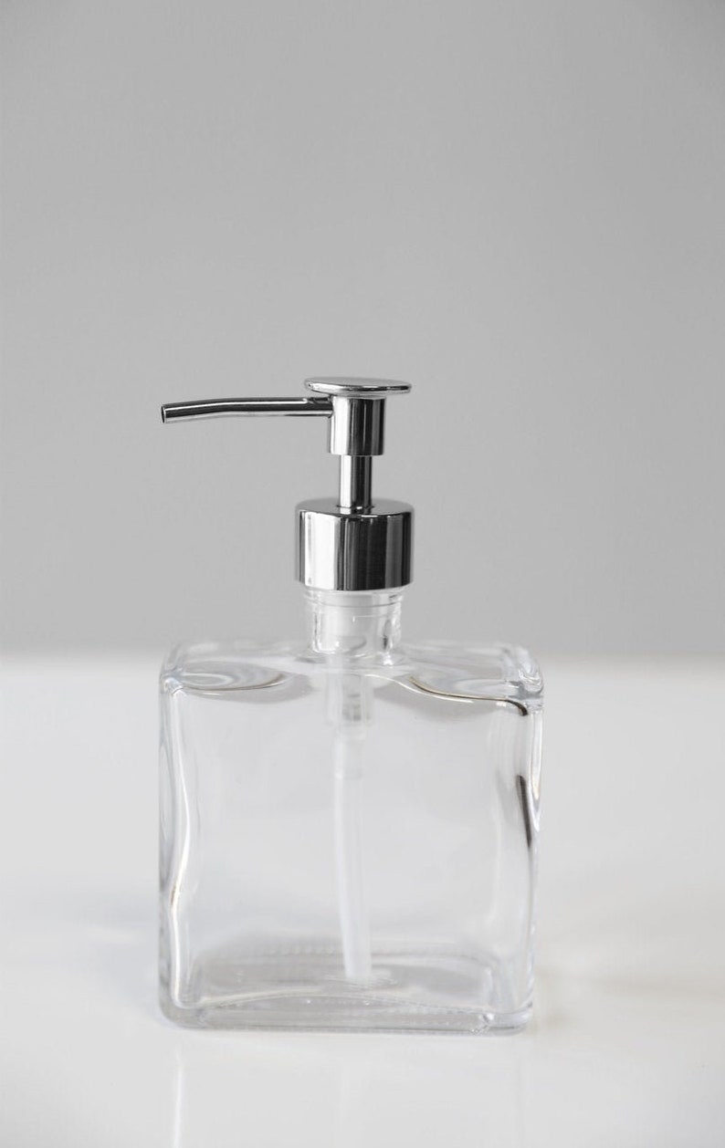 May include: A clear glass soap dispenser with a chrome pump top. The dispenser has a square shape and is sitting on a white surface.