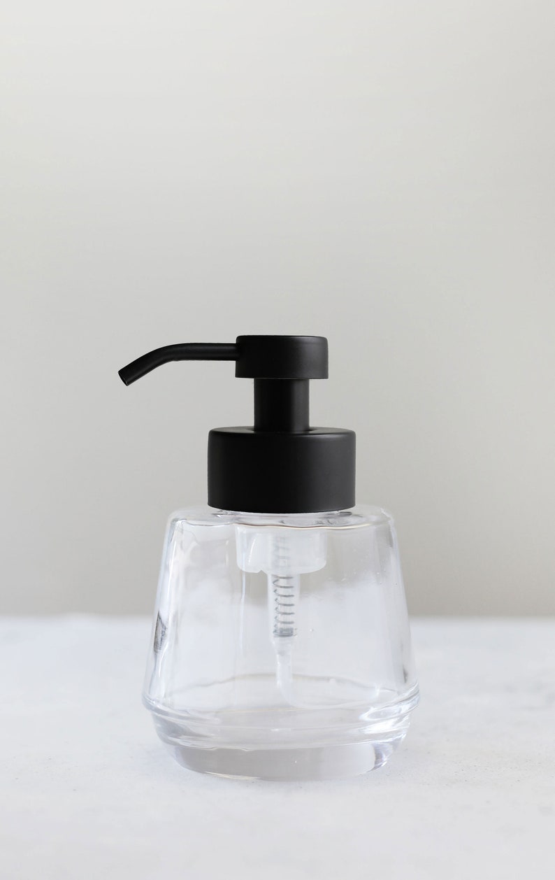 K&ouml;nnte beinhalten: Ein transparenter Seifenspender aus Glas mit einer schwarzen Pumpe. Der Spender hat ein strukturiertes, facettiertes Design.