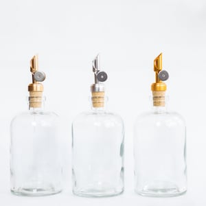 May include: Three clear glass bottles with cork stoppers and metal pour spouts. The spouts are gold, silver, and gold.