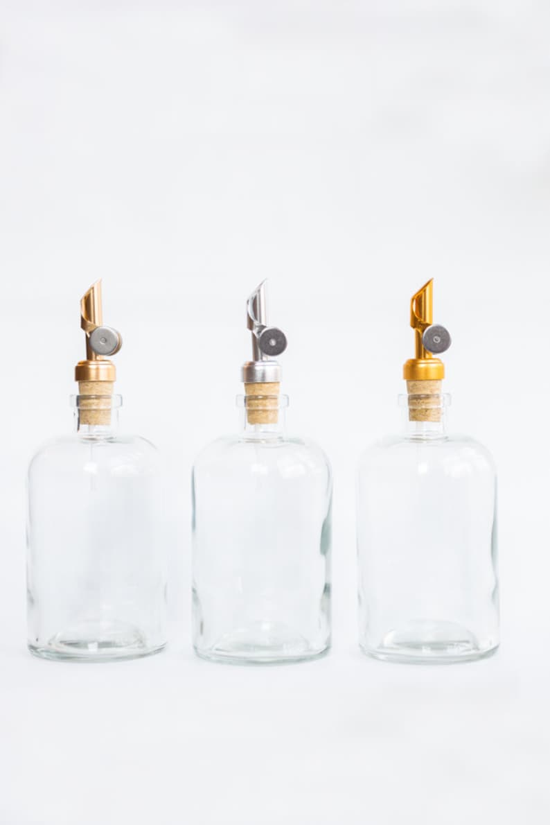 May include: Three clear glass bottles with cork stoppers and metal pour spouts. The spouts are gold, silver, and gold.