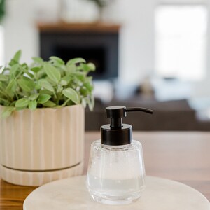 K&ouml;nnte beinhalten: Ein durchsichtiger Seifenspender aus Glas mit einer schwarzen Pumpe und einer wei&szlig;en Marmor-Seifenschale. Der Spender ist mit einer schaumigen Fl&uuml;ssigkeit gef&uuml;llt.