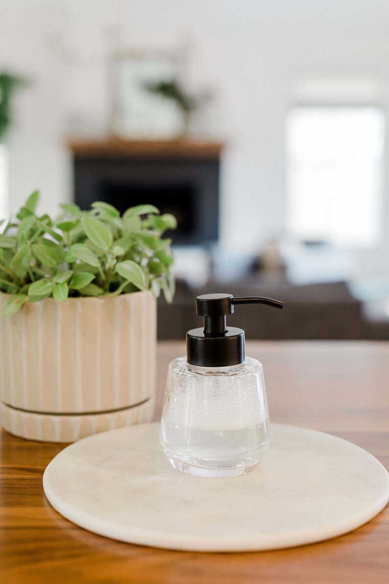 K&ouml;nnte beinhalten: Ein durchsichtiger Seifenspender aus Glas mit einer schwarzen Pumpe und einer wei&szlig;en Marmor-Seifenschale. Der Spender ist mit einer schaumigen Fl&uuml;ssigkeit gef&uuml;llt.