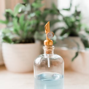 May include: A clear glass bottle with a blue liquid inside and a gold metal pump top. The bottle is sitting on a white surface.