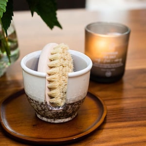 May include: A white ceramic brush holder with a brown speckled glaze. A wooden handled brush with natural bristles is in the holder. The holder is on a wooden tray.