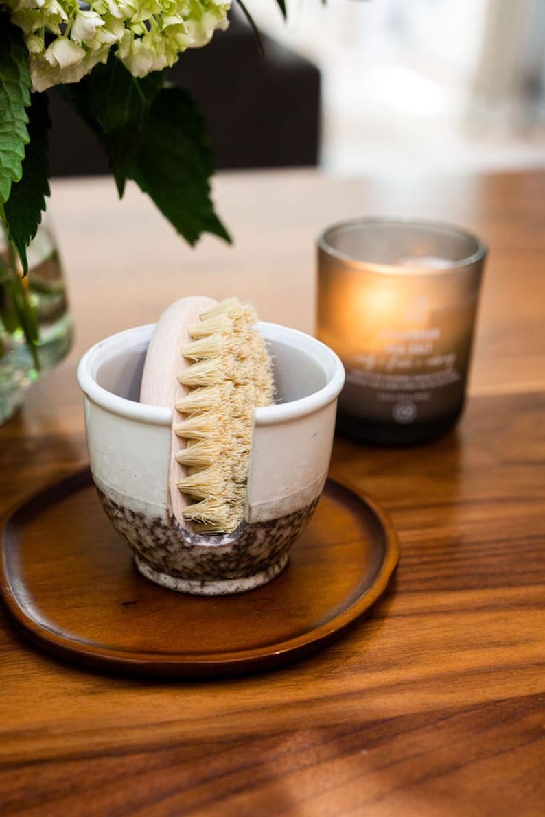 May include: A white ceramic brush holder with a brown speckled glaze. A wooden handled brush with natural bristles is in the holder. The holder is on a wooden tray.