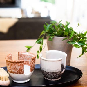 May include: Two ceramic dish soap holders on a black tray. One holder is brown and white with speckles, the other is white with black speckles. A wooden brush is in the foreground.