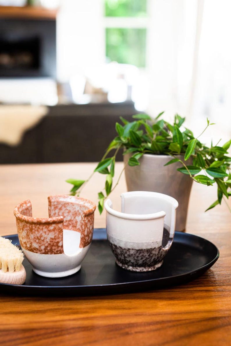 May include: Two ceramic dish soap holders on a black tray. One holder is brown and white with speckles, the other is white with black speckles. A wooden brush is in the foreground.