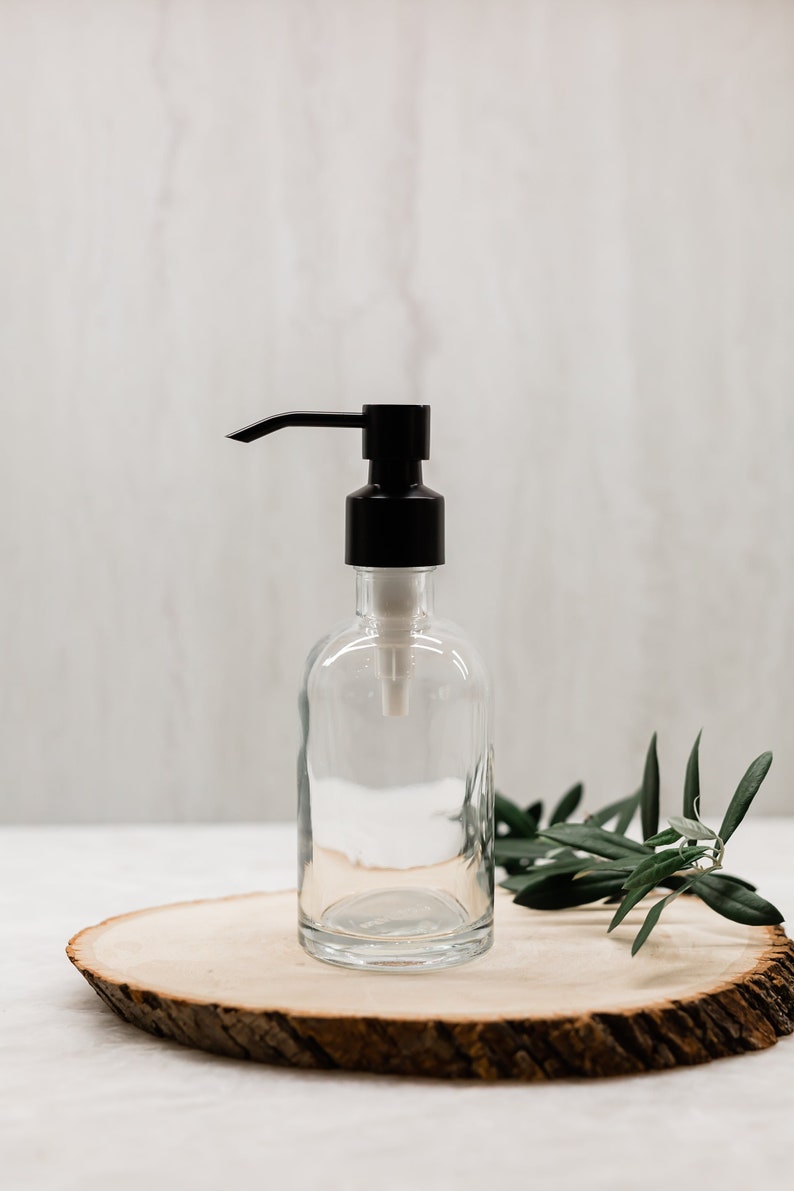 May include: A clear glass bottle with a black pump dispenser, sitting on a wooden slice with green leaves.