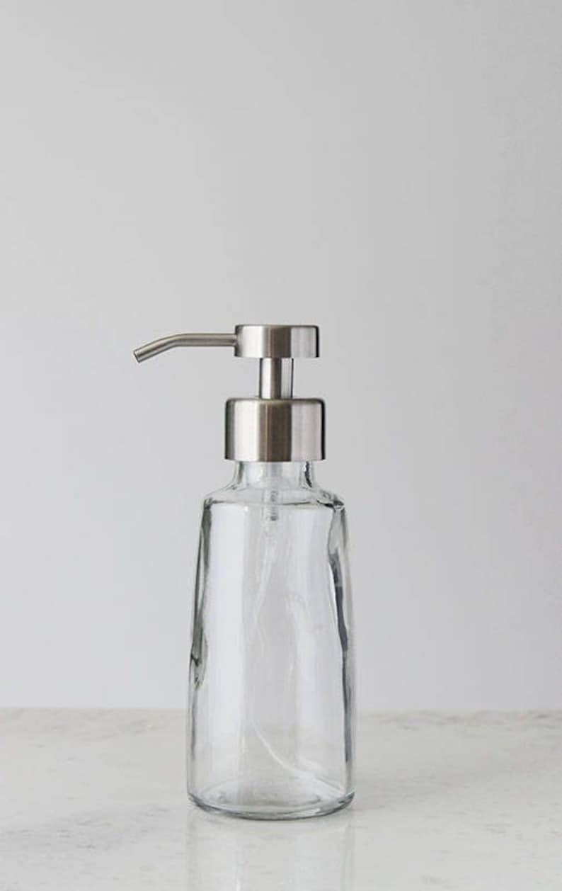 May include: A clear glass soap dispenser with a stainless steel pump. The dispenser has a simple, modern design.