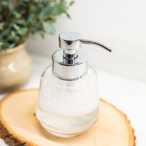 Pu&ograve; includere: Un dispenser di sapone in vetro trasparente con una pompa argentata e un liquido schiumoso bianco all'interno. Il dispenser &egrave; appoggiato su una fetta di legno.