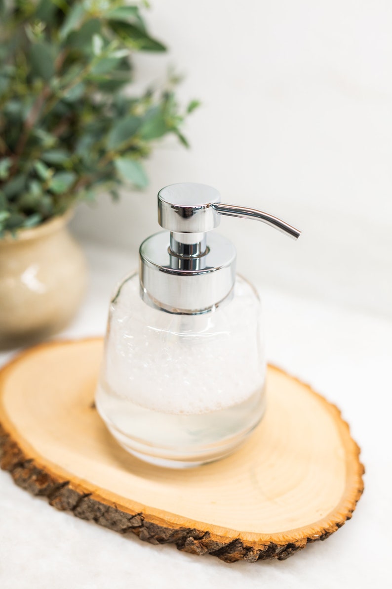 Pu&ograve; includere: Un dispenser di sapone in vetro trasparente con una pompa argentata e un liquido schiumoso bianco all'interno. Il dispenser &egrave; appoggiato su una fetta di legno.