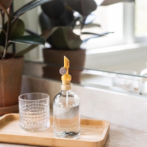May include: A clear glass bottle with a gold pump dispenser and a cork stopper sits on a wooden tray with a clear glass tumbler. The bottle and tumbler are on a white countertop.