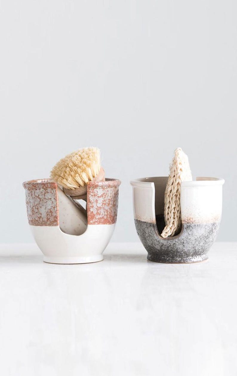 May include: Two ceramic dish soap and sponge holders with a speckled glaze. One holder is white with a brown band and holds a brush. The other holder is white with a black band and holds a sponge.