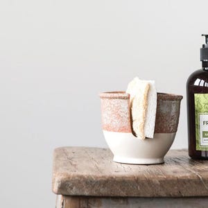 May include: A white ceramic sponge holder with a brown glaze and a cutout for holding a sponge. The holder is sitting on a wooden table next to a bottle of hand soap.