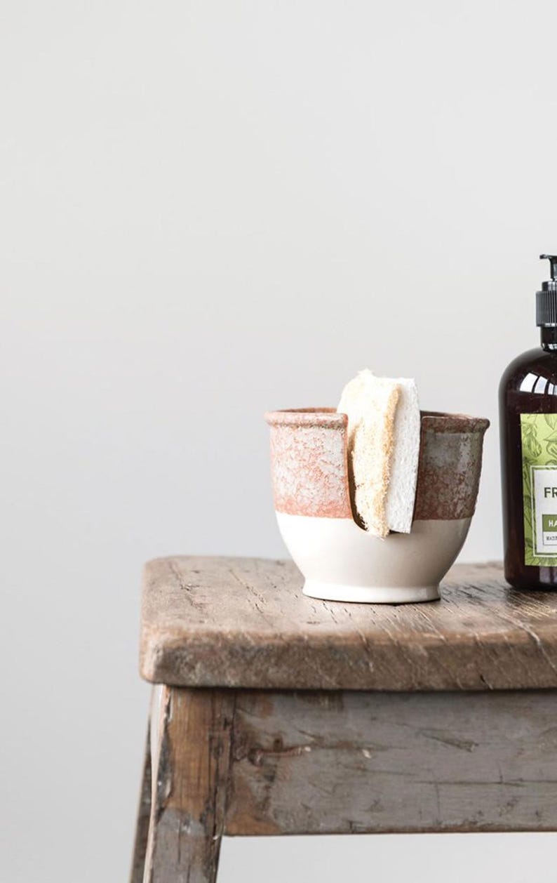 May include: A white ceramic sponge holder with a brown glaze and a cutout for holding a sponge. The holder is sitting on a wooden table next to a bottle of hand soap.