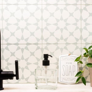 May include: A clear glass soap dispenser with a black pump sits on a white countertop in front of a white tiled backsplash with a geometric pattern. A white sign with the text "USE SOAP" is to the right of the dispenser.