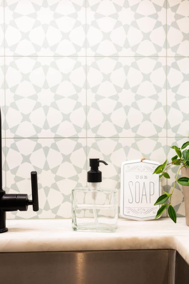 May include: A clear glass soap dispenser with a black pump sits on a white countertop in front of a white tiled backsplash with a geometric pattern. A white sign with the text "USE SOAP" is to the right of the dispenser.