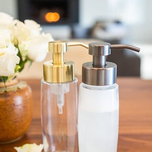 May include: Two glass soap dispensers with pumps. The dispenser on the left has a gold pump and the dispenser on the right has a dark bronze pump. Both dispensers are filled with a white liquid.