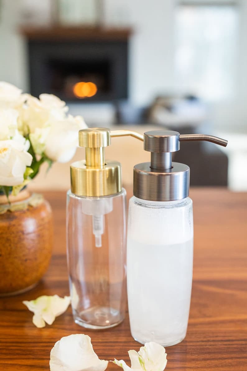 May include: Two glass soap dispensers with pumps. The dispenser on the left has a gold pump and the dispenser on the right has a dark bronze pump. Both dispensers are filled with a white liquid.