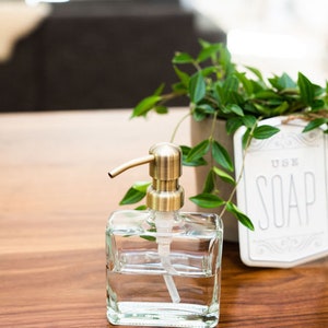 May include: A clear glass soap dispenser with a gold pump top. The dispenser is filled with liquid soap and is sitting on a wooden surface. A small potted plant and a sign that says "Use Soap" are in the background.