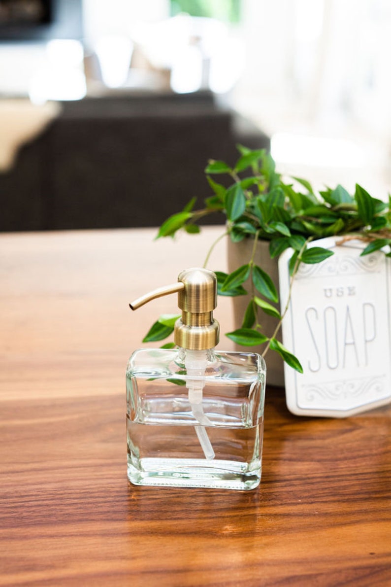 May include: A clear glass soap dispenser with a gold pump top. The dispenser is filled with liquid soap and is sitting on a wooden surface. A small potted plant and a sign that says "Use Soap" are in the background.
