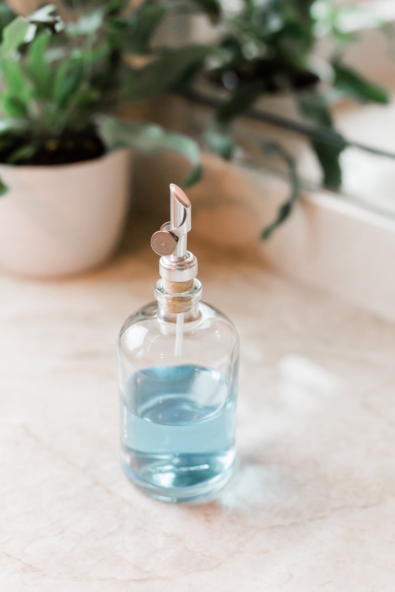 May include: A clear glass bottle with a silver pump dispenser filled with light blue liquid. The bottle is sitting on a white marble surface.