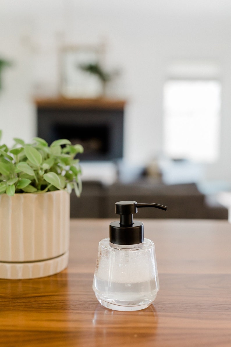 K&ouml;nnte beinhalten: Eine klare Glaspumpflasche mit schwarzem Pumpkopf, gef&uuml;llt mit einer klaren Fl&uuml;ssigkeit, steht auf einem Holztisch. Im Hintergrund befindet sich eine Topfpflanze.